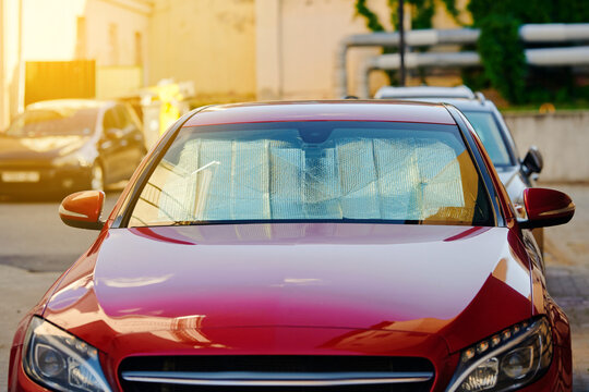 Front view of modern red car parked in an outdoor urban lot with reflective silver sunshade placed inside the windshield for heat and uv protection during bright sunny day. Sun protector shield