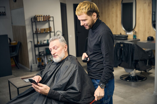 Mature man using smartphone while sitting in barbershop, talking with barber