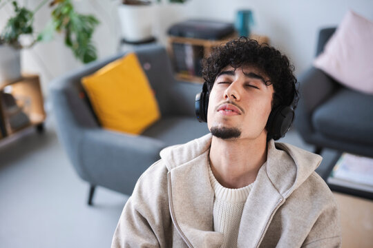 A young man with curly hair listens to music with headphones on, eyes closed, in a relaxed indoor setting.