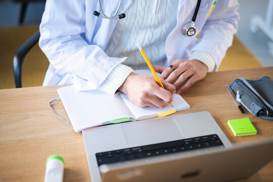 A medical professional in a white coat writes in a notebook at a desk with a laptop and medical equipment.