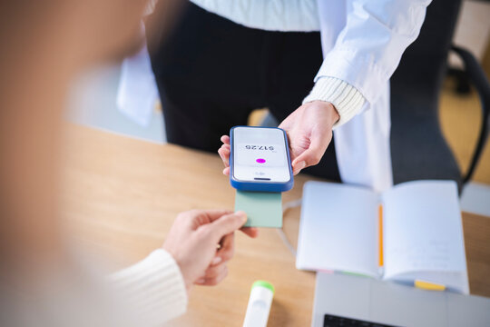 A person in a white coat holds a smartphone displaying a payment amount, while another person holds a card to complete a transaction.