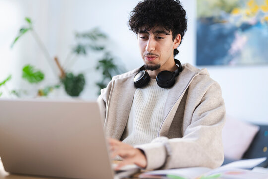 A young man with curly hair wearing headphones around his neck works on his laptop at a desk, surrounded by plants and art.