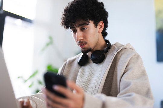A young man with curly hair wears headphones around his neck while looking at his phone. He is seated indoors, with a laptop in front of him.