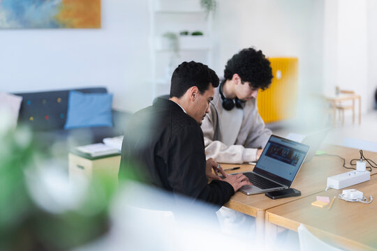 Two young men collaborate on a project using laptops at a wooden table, surrounded by office supplies and a comfortable seating area in the background.