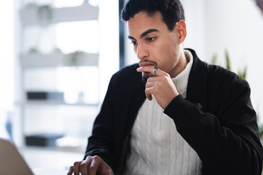 A thoughtful man in a black jacket and white sweater looks down at his laptop, his finger touching his chin as he concentrates on his work.