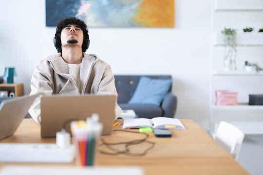 A young man with headphones on leans back in his chair, eyes closed, enjoying music while working on his laptop at a desk.