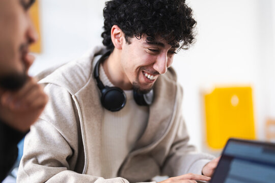 A young man with curly hair smiles while working on a laptop, with headphones around his neck, in a bright, modern setting.