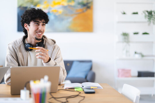 A young man with curly hair sits at a desk with a laptop, holding a pencil and wearing headphones. He is engaged in his work or studies.