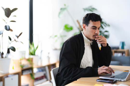 A young man sits at a desk, focused on his laptop. He is dressed in a black jacket over a cream cable-knit sweater.
