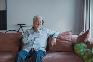 Fototapeta premium Elderly man sitting comfortably on a sofa at home, smiling gently while looking out the window, enjoying a calm and joyful moment of reflection, feeling content and peaceful during his daily routine