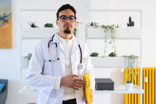 A male doctor wearing a white coat and stethoscope holds a yellow folder. He stands in a modern office with plants and shelves in the background.
