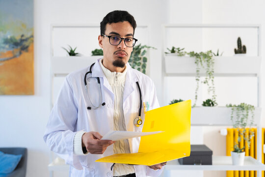 A male doctor in a white coat and stethoscope holds a yellow folder and papers, reviewing medical information in an office setting.
