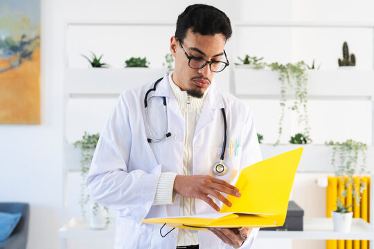 A male doctor in a white coat and stethoscope reviews documents in a yellow folder, focusing on patient information.