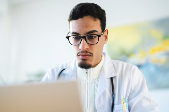 A focused medical professional wearing glasses and a lab coat works on a laptop, reviewing patient information.