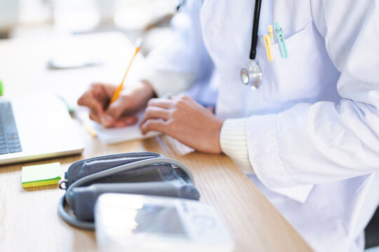 A medical professional in a white coat writes notes at a desk with a stethoscope, laptop, and blood pressure monitor.