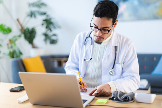 A doctor in a white coat and stethoscope is writing on a notepad while looking at a laptop.