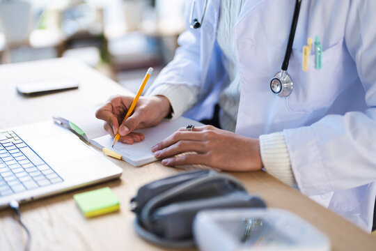 A medical professional in a white coat writes in a notebook with a pencil, with a stethoscope around their neck and a laptop on the desk.