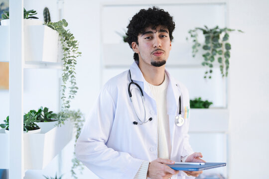 A young male doctor in a white coat and stethoscope holds a tablet, looking directly at the camera in a modern office setting with plants.