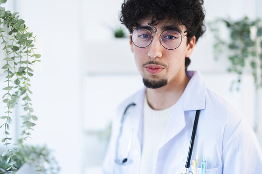 A young male doctor wearing glasses and a white coat with a stethoscope around his neck looks thoughtfully into the camera.