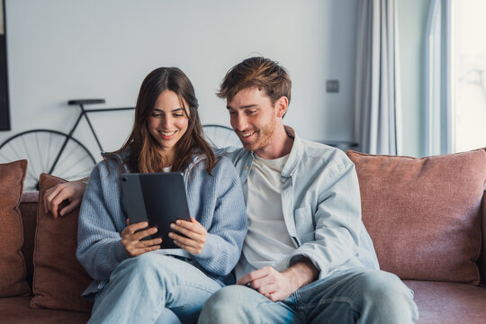 Happy young couple relaxing on cozy couch, using computer tablet together, smiling beautiful woman and man hugging, looking at device screen, watching movie or video, shopping online, browsing apps