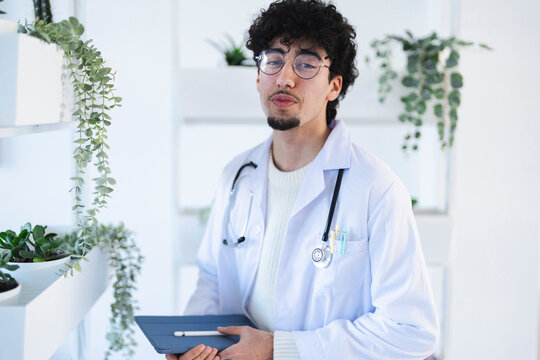 A young male doctor in a white coat and stethoscope holds a tablet and stylus, standing near shelves with plants.