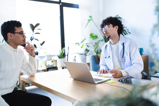 A doctor in a white coat consults with a patient, taking notes on a pad while a laptop sits on the desk.