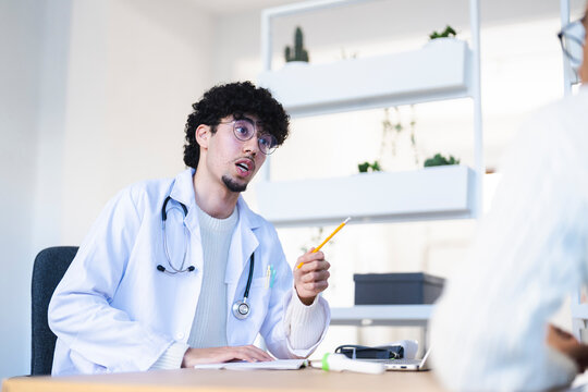 A doctor in a white coat with a stethoscope is talking to a patient, holding a pencil and looking at notes on a desk.