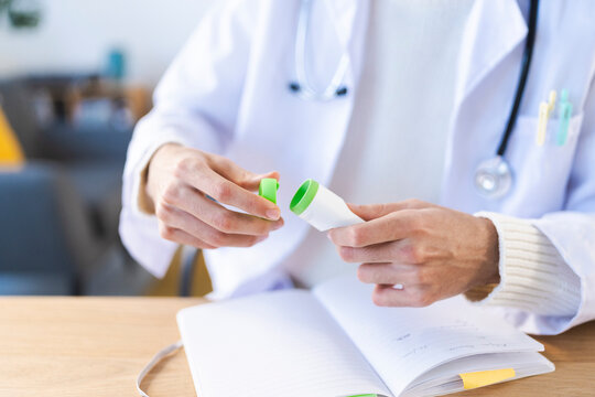 A medical professional in a white coat opens a small container of medication, with a notebook open on the desk.