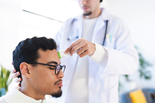 A medical professional in a white coat uses a thermometer to check a patient's temperature on their forehead during a health checkup.
