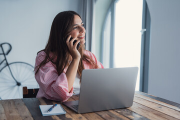 Smiling young lady working at home office desk by laptop engaged in phone conversation with client...