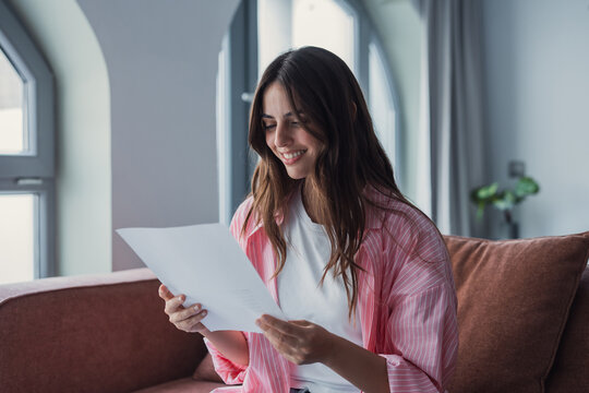 Smiling attractive young woman indoor sit on sofa at home reading letter. Pleased female holds in hands sheet pleased by good news written in official document. Bank notice, happy client concept