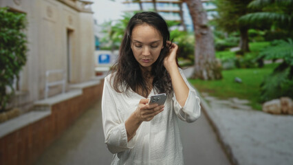 Fototapeta premium Woman tapping smartphone with both hands, looking down while walking on a narrow street beside a building, surrounded by trees and a walkway; contemplation.