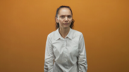 Fototapeta premium Woman in white shirt frowning with furrowed brow and tilted head against orange studio backdrop, hands relaxed at sides; irritation.