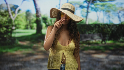 Woman in straw hat and yellow crochet top stands with finger to lips in forest, wearing yellow sunglasses and jeans  hush. © Krakenimages.com