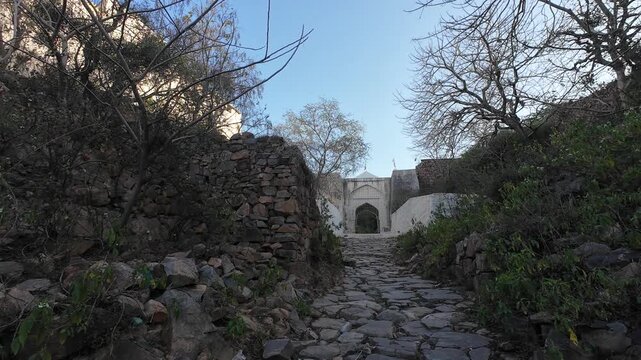 A historic white-painted main gate and stone pathway leading to a religious shrine situated on a rugged hill within the Aravalli Range in Nuh district