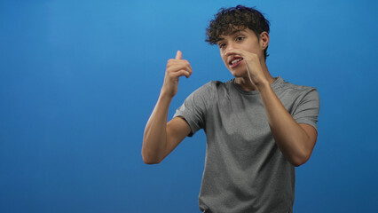 Teenage boy making animated hand gestures and touching his mouth while speaking in a blue studio, wearing a grey t shirt and looking engaged  expressive. © Krakenimages.com