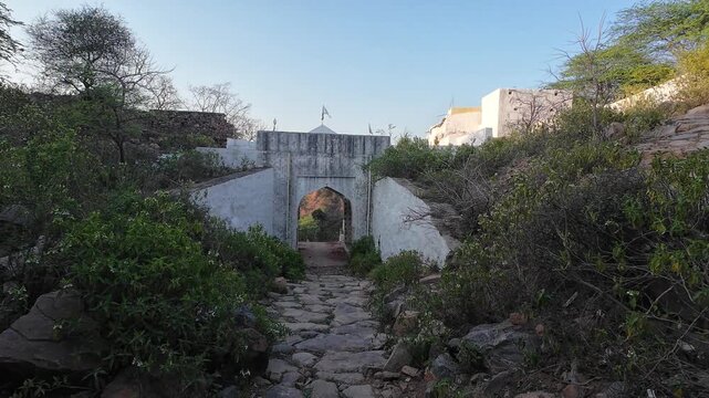 A historic white-painted main gate and stone pathway leading to a religious shrine situated on a rugged hill within the Aravalli Range in Nuh district