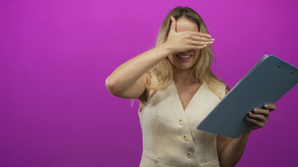 Fototapeta premium Young blonde woman holding blue clipboard covers eyes with hand while smiling and turning slightly in purple studio; playful shyness.