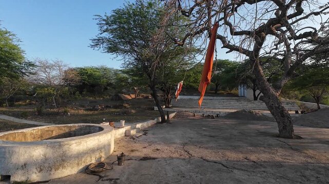 An ancient Hindu temple shrine situated on a rocky hill in the Aravalli range featuring traditional white-washed architecture and old twisted trees under a clear sky.