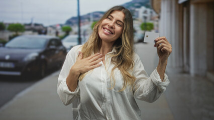Fototapeta premium Middle aged woman holding creditcard in hand on street, smiling with open mouth and waving the card toward camera; consumer confidence joy.
