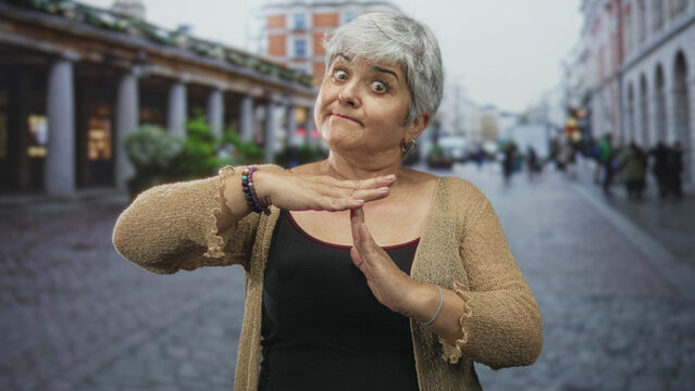 Senior woman with grey hair and bracelets shows hands forming timeout sign on cobbled pedestrian street by colonnade and shops; firm disapproval.