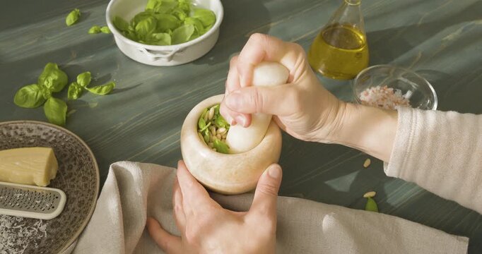 Hands grinding pine nuts and basil in marble mortar for Genovese pesto. Close-up of person using stone pestle to crush ingredients for traditional Italian sauce on wooden table. Making authentic pesto