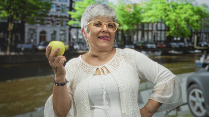 Fototapeta premium Woman holding a green apple in hand while examining it on a street canal lined with buildings; quiet contemplation.