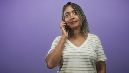 Fototapeta premium Woman holding smartphone to ear while checking messages and then smiling in studio purple backdrop; casual conversation content.