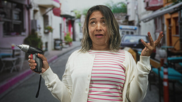 Hispanic middle age woman holding a cordless screwdriver and covering her face with hand in a narrow street cafe, striped shirt and cardigan visible; diy frustration.