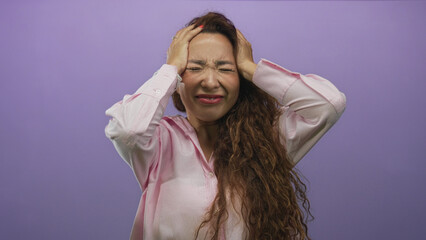 Fototapeta premium Young caucasian woman with long curly hair, hands raised and holding her head in a purple studio; frustration stress.