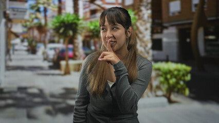 Fototapeta premium Woman points finger to lips for silence on a sunny urban street lined with palm trees and blurred storefronts, looking at camera; playful secret.