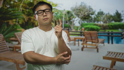 Young chinese man in white shirt stands pointing finger at crystal pool beside resort building under leafy palms  curiosity. © Krakenimages.com