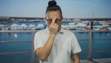 Fototapeta premium Man pinching bridge of nose with two fingers on street beside marina boats and metal railing, wearing white polo and glasses; disgust.