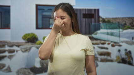 Woman pinching nose in yellow t shirt at building entrance of house outdoors near glass railing;...